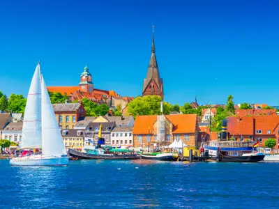 Cityscape of Flensburg. Panorama of a small European town in Northern Germany. A sailboat is floating in a harbour along the coastline with old architecture, ships and landmarks on the background