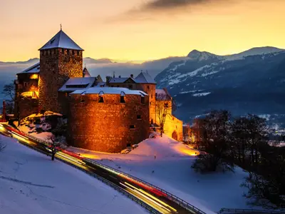 Vaduz, Liechtenstein. Illuminated castle of Vaduz at sunset - popular landmark at night, with car traffic lights and sunset sky, mountains at the background