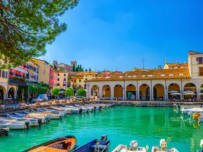 Old harbour Porto Vecchio with motor boats on turquoise water, green trees and traditional buildings in historical centre of Desenzano del Garda town, blue sky background, Lombardy, Northern Italy